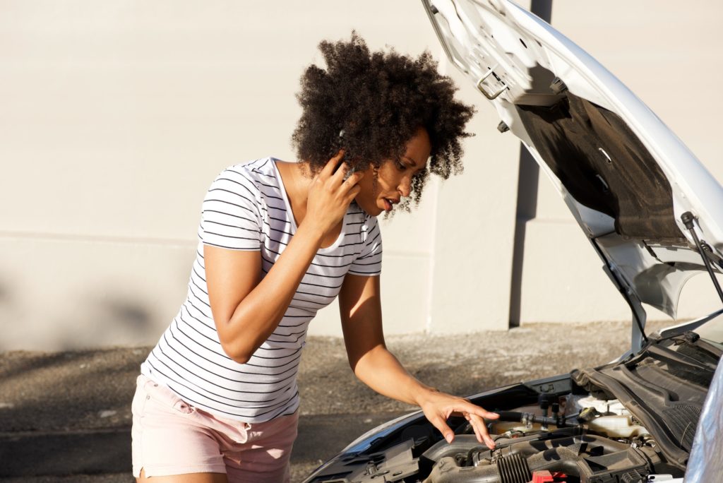 young african woman standing by broken down car parked on the road and calling for assistance